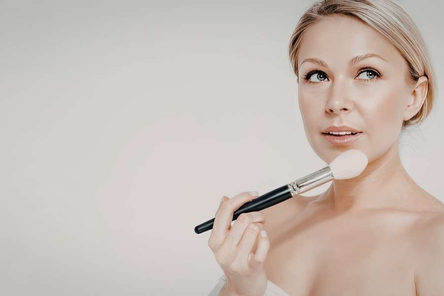 Femme blonde appliquant un pinceau de maquillage sur son visage dans le cadre de soins du visage spécifiques.