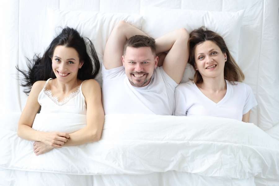 Un homme et deux femmes souriants allongés ensemble sous une couette blanche dans un lit lumineux.